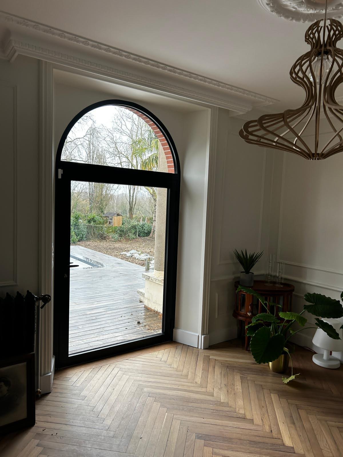 Porte-fenêtre cintrée en aluminium noir posée dans une pièce de vie avec parquet en chevrons, par Bonno Ouvertures.