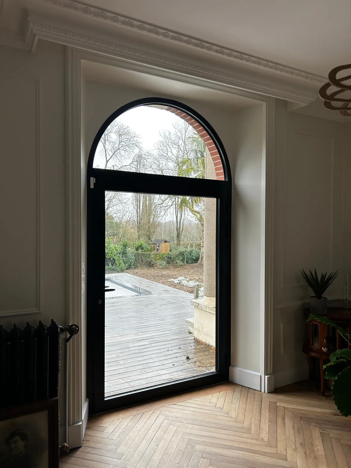 Porte d’entrée cintrée en aluminium noir avec vitrage, donnant sur une terrasse boisée, posée par Bonno Ouvertures.
